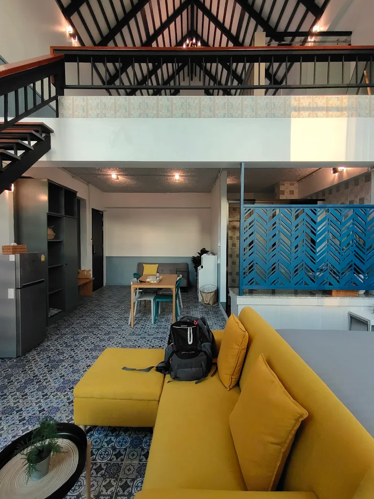 Wide interior view of a big villa duplex, with a spiral staircase to the mezzanine, a mustard sofa, and a kitchenette.